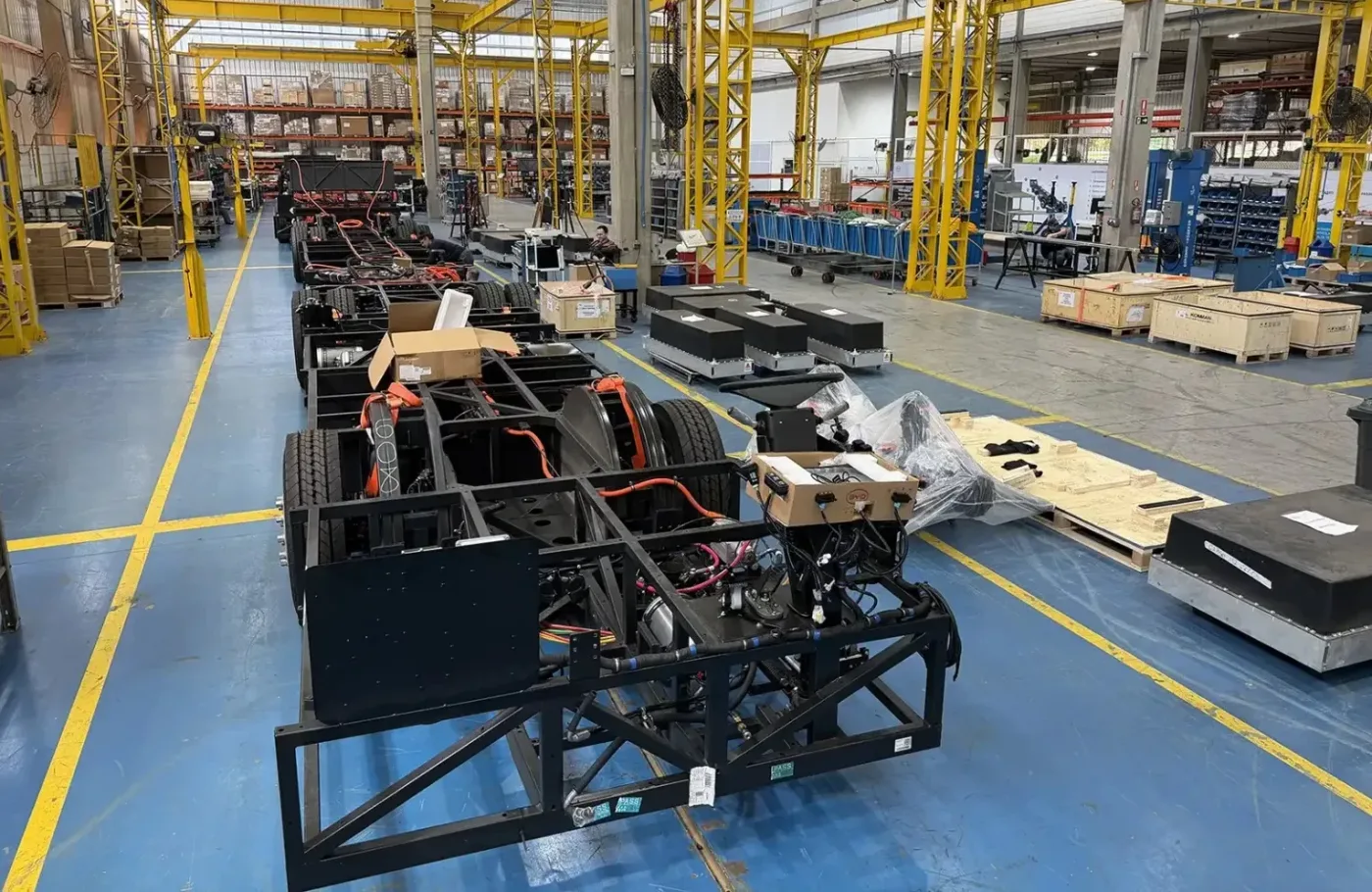 Chassis de ônibus elétricos em linha de montagem industrial, com estruturas metálicas pretas, pneus e fiação laranja. Caixas de papelão e madeira, além de comp