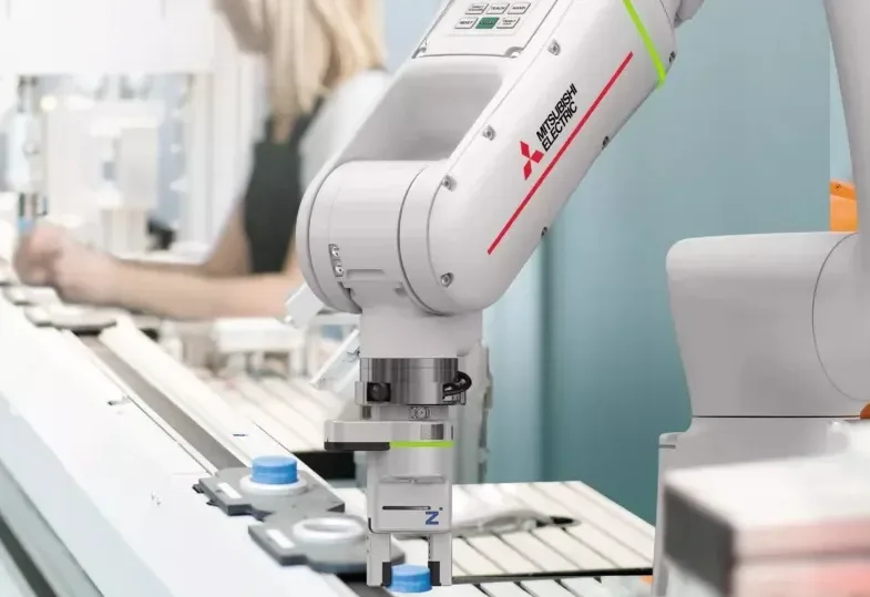 Braço robótico branco da Mitsubishi Electric com detalhes vermelhos e verdes, manipulando uma peça com tampa azul em uma linha de montagem. Ao fundo, uma pesso