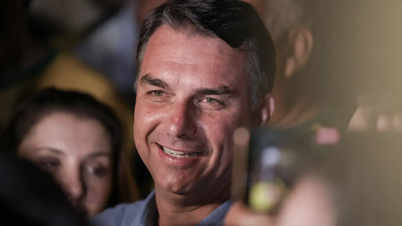 Homem de meia-idade, cabelo escuro e camisa azul, sorri amplamente mostrando os dentes, em meio a uma multidão desfocada. Ele olha para a esquerda, com luz sua
