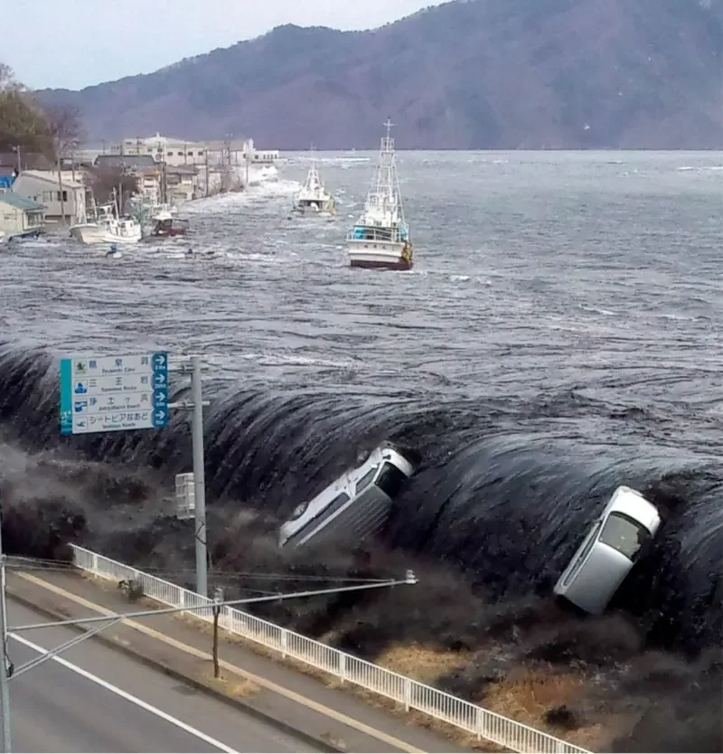 Uma grande onda escura de tsunami avança sobre uma cidade costeira, inundando ruas e prédios, enquanto dois carros de cor clara são arrastados pela força da ág