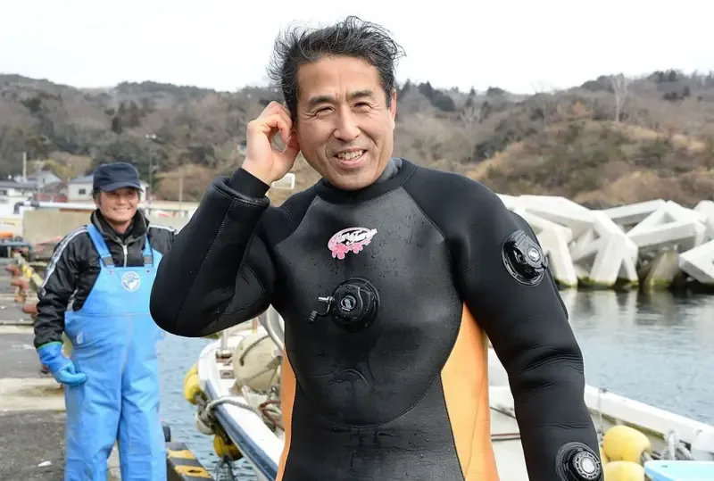 Um homem de roupa de mergulho preta aparece em primeiro plano, em um cais, ao lado de um pequeno barco, com uma das mãos erguida junto à cabeça e gotas de água