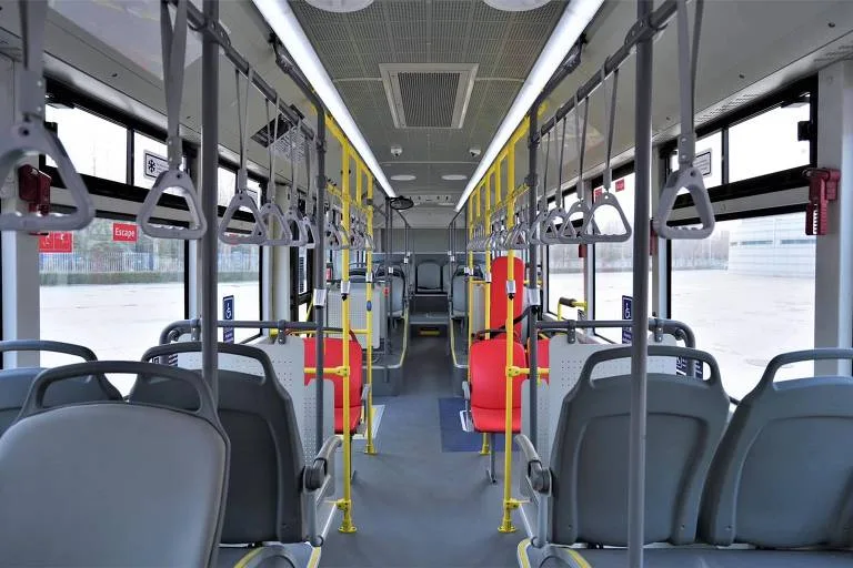 Interior de ônibus urbano com fileiras de assentos cinza e vermelhos, barras amarelas para apoio e alças de mão suspensas. Janelas amplas permitem visão extern